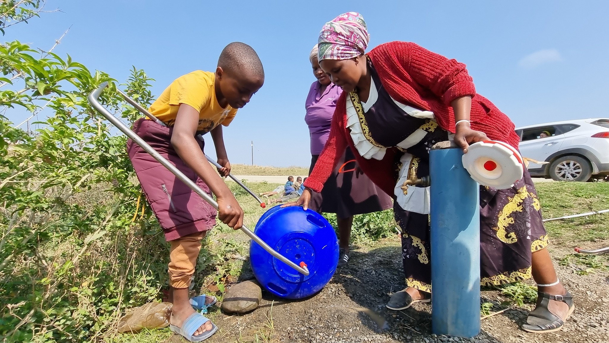 A child assists a woman at a water source, filling a blue container while another adult observes. This scene highlights community engagement in water access.