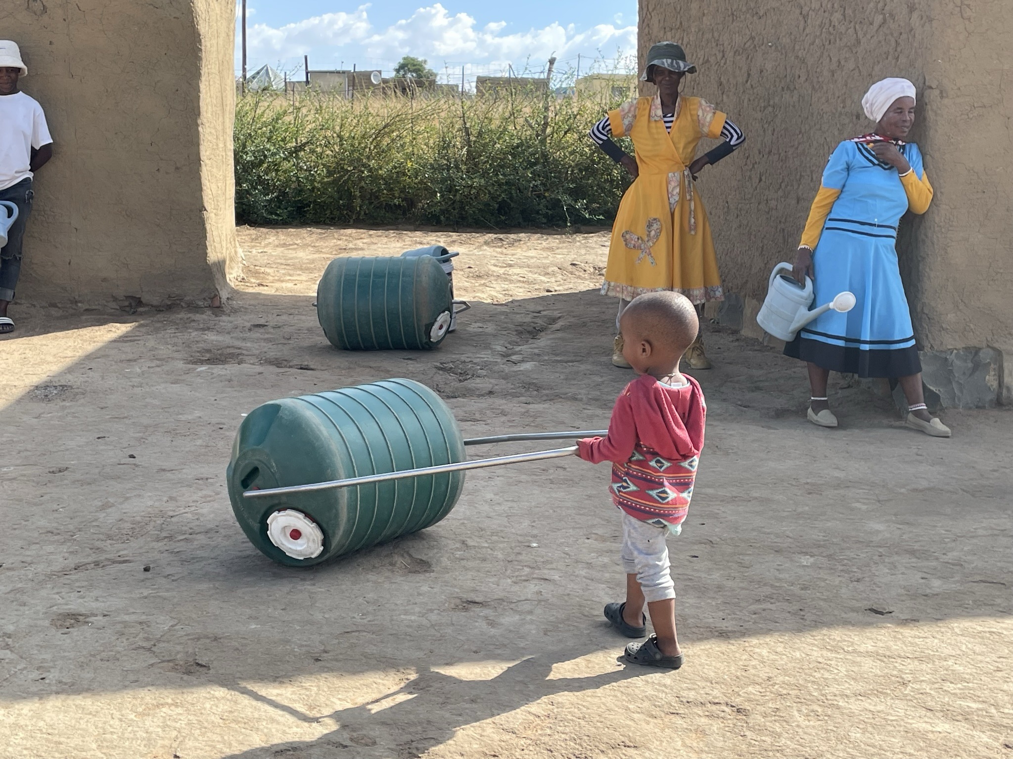 A child pulls a large water container with a handle while two women in traditional attire observe nearby, set against a rural backdrop.