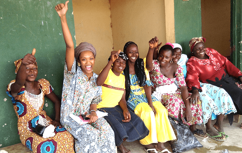 Seven young women sitting against a wall, smiling and cheering with thumbs up, celebrate together, showcasing joy and community spirit.