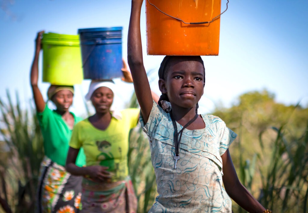 Young girl carrying an orange bucket on her head, surrounded by women with colorful containers, illustrating daily water collection in rural life.