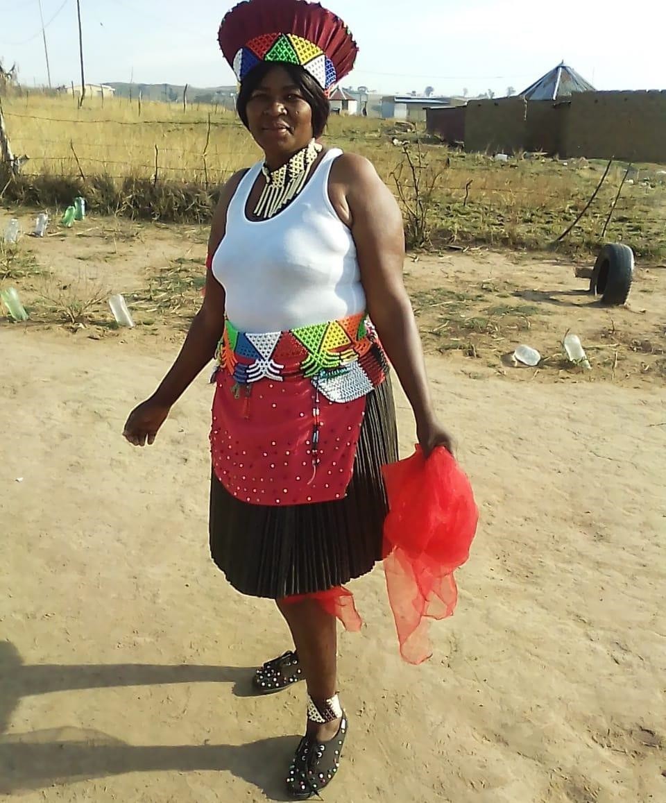 A woman stands in traditional attire, adorned with colorful beaded jewelry and a vibrant headdress, set against a rural backdrop.