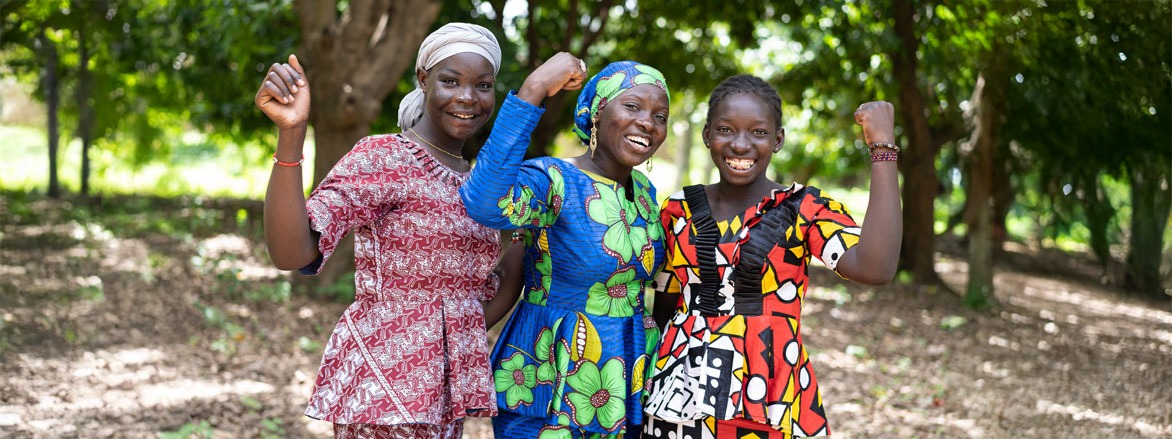 Three women in vibrant, African-patterned dresses celebrate joyfully outdoors, showcasing empowerment and community spirit among them.