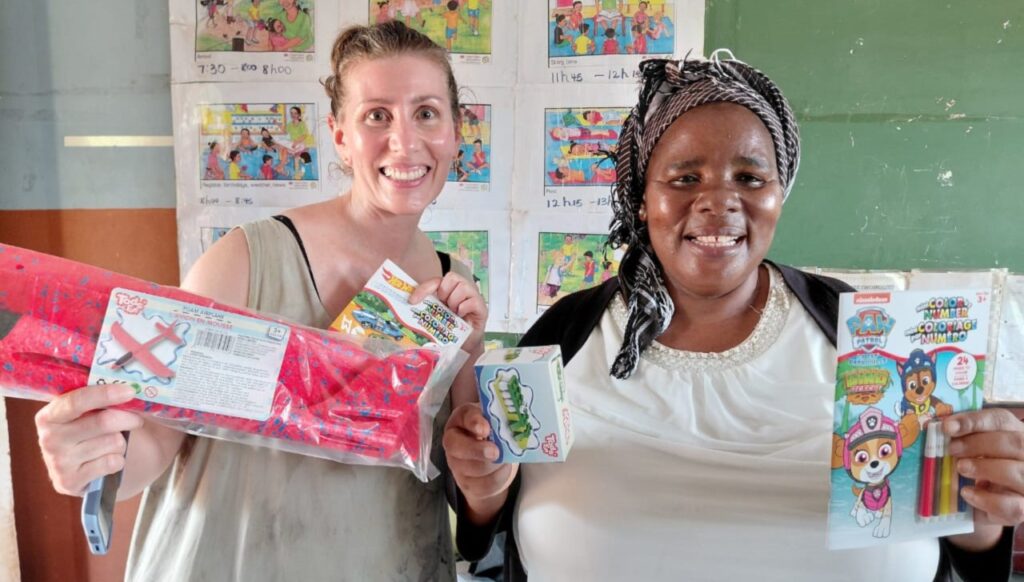 Two women smile while holding educational toys and activity books in a classroom, showcasing materials for children's learning and play.