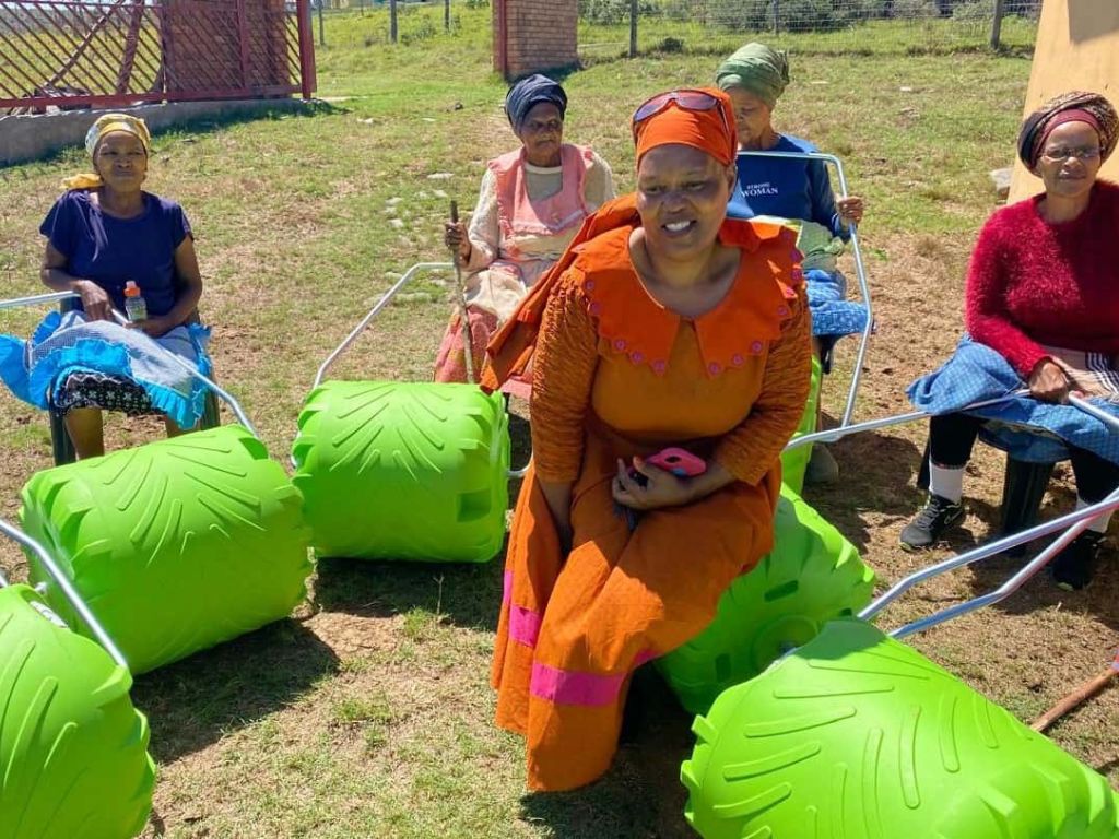 A group of five women, dressed in vibrant traditional attire, sits outdoors on bright green chairs. They engage in conversation, highlighting community and cultural connection.