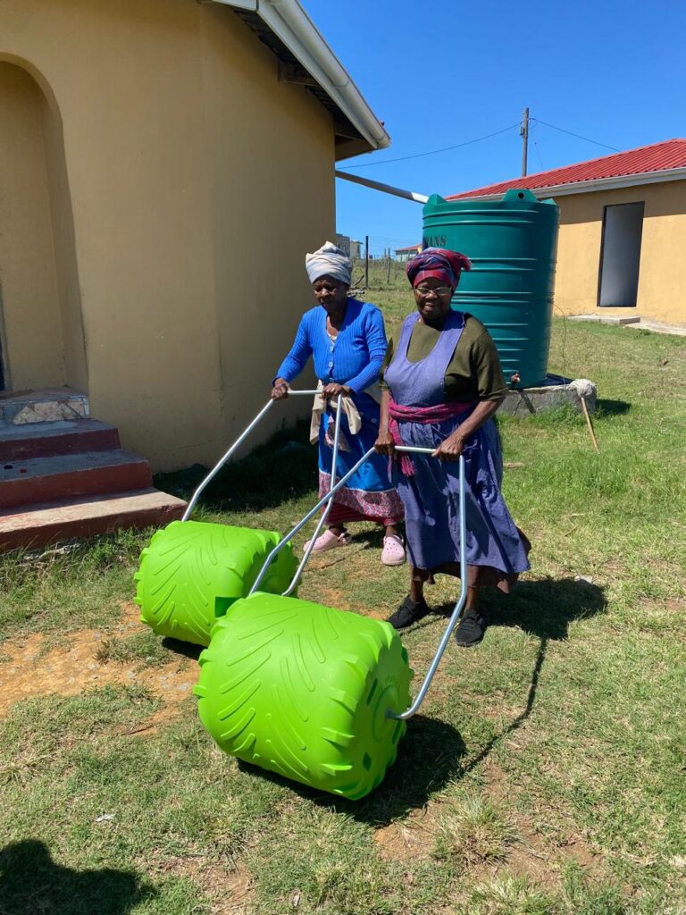Two women in traditional attire pull unique green water transport devices across a grassy field, highlighting innovative solutions for rural communities.