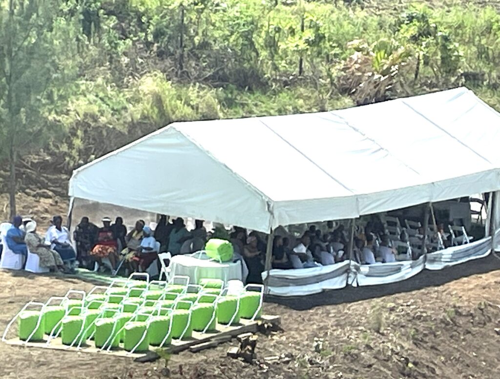 A white tent hosts an audience sitting on folding chairs, surrounded by greenery. Bright green containers are arranged in front, suggesting an event setup.