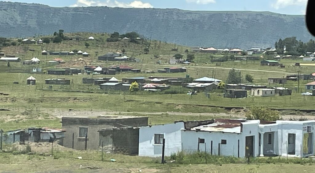 Rural landscape with sparse housing, featuring a mix of traditional huts and modern structures against a backdrop of green hills and blue sky.
