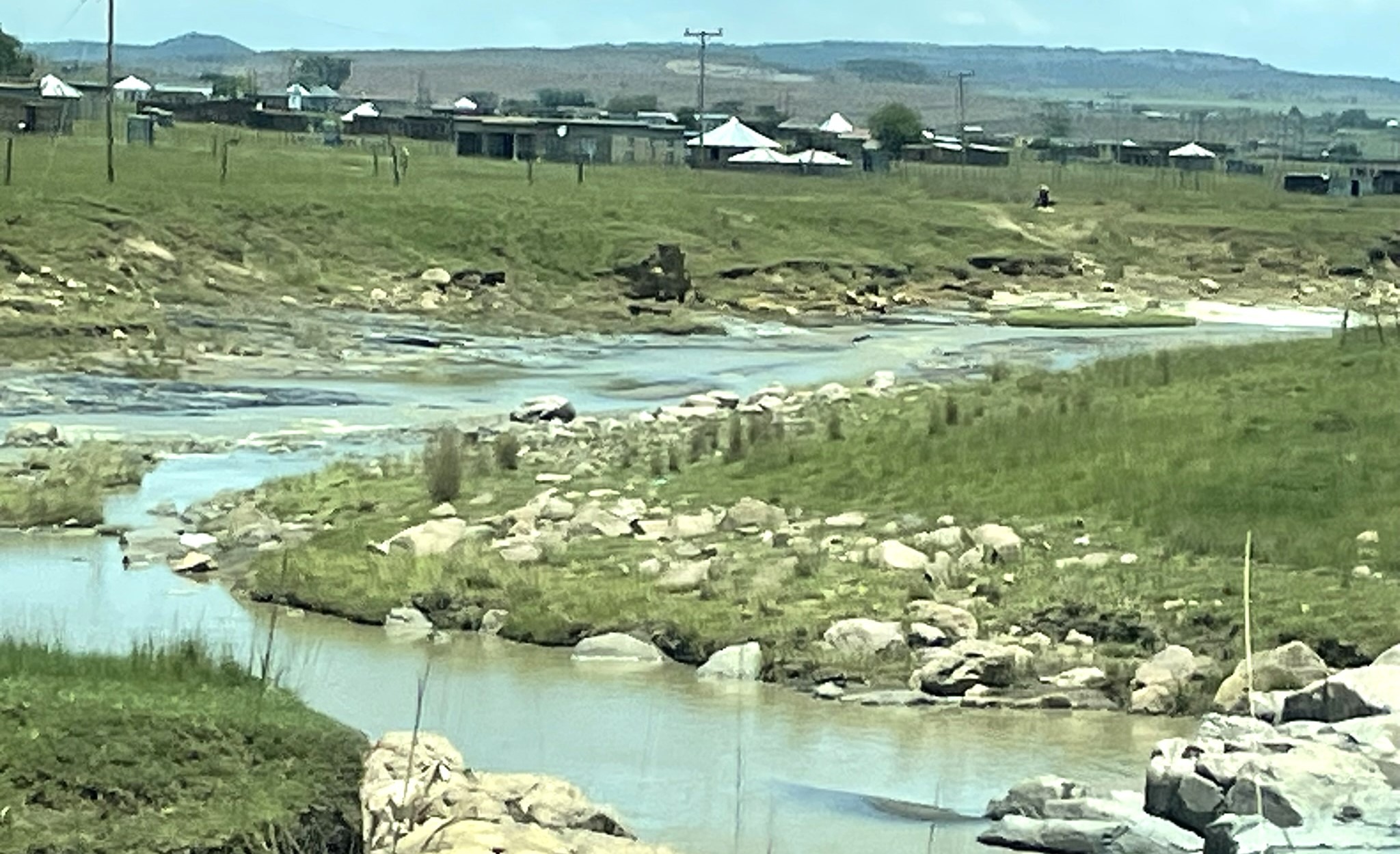 A winding river flows through a grassy landscape dotted with rocks, framed by distant hills and clusters of structures.