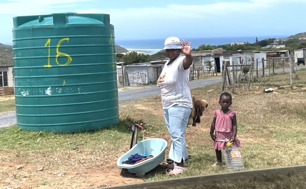 A woman waves while standing near a water tank and a basin of clothes, with a child beside her, in a rural community setting.
