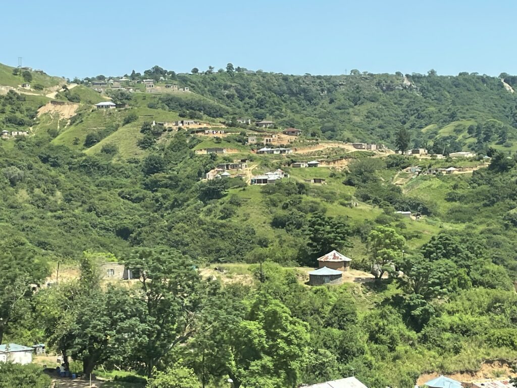 Lush green hills dotted with small houses under a clear blue sky. The scene depicts rural living amidst a natural landscape.