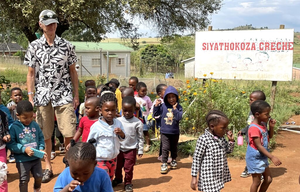 A diverse group of young children walks past the Siyathokoza Creche sign, accompanied by an adult in a floral shirt. The scene captures a vibrant community atmosphere.