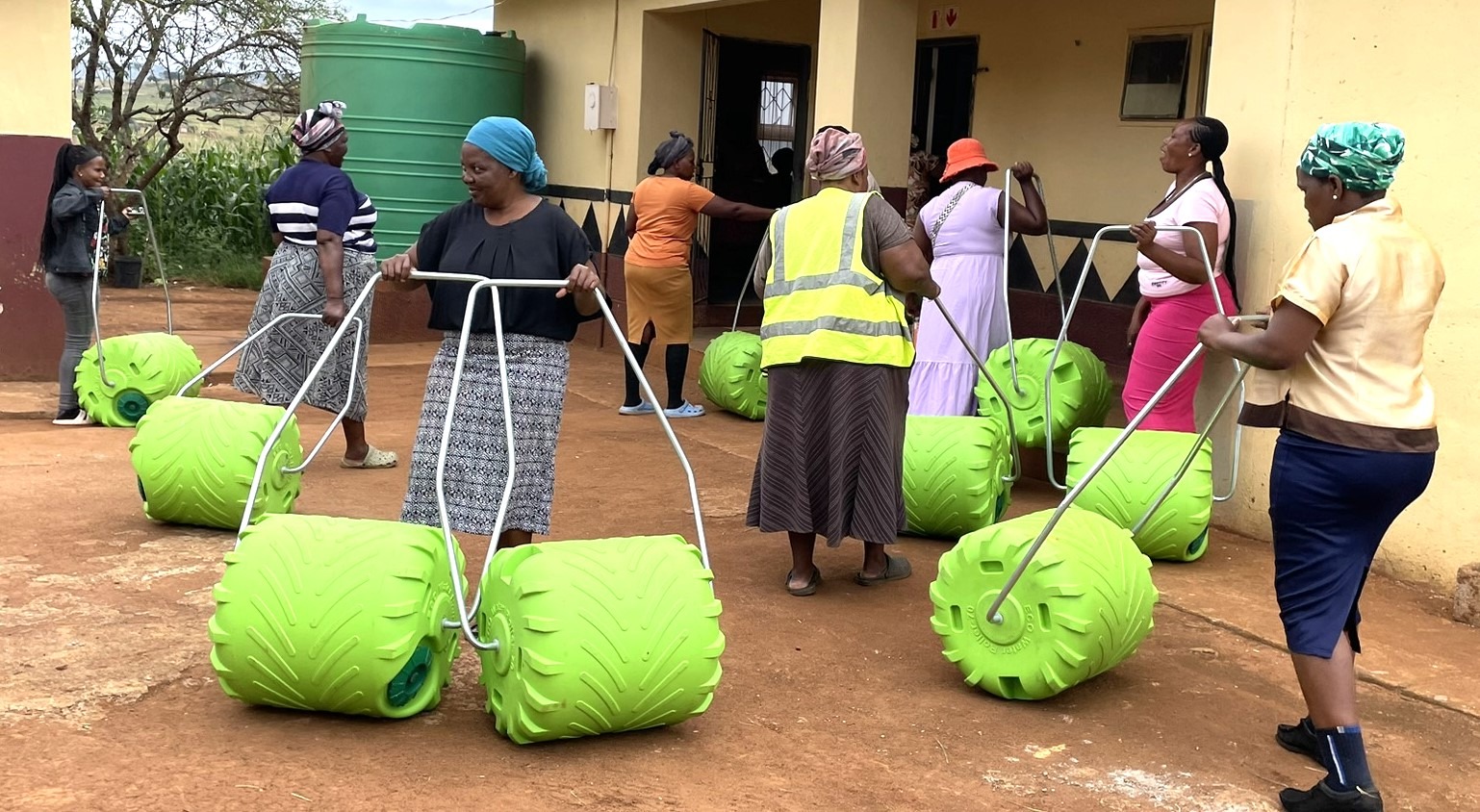 Women transport green, wheeled water containers outside a building, demonstrating community efforts to improve access to water resources.