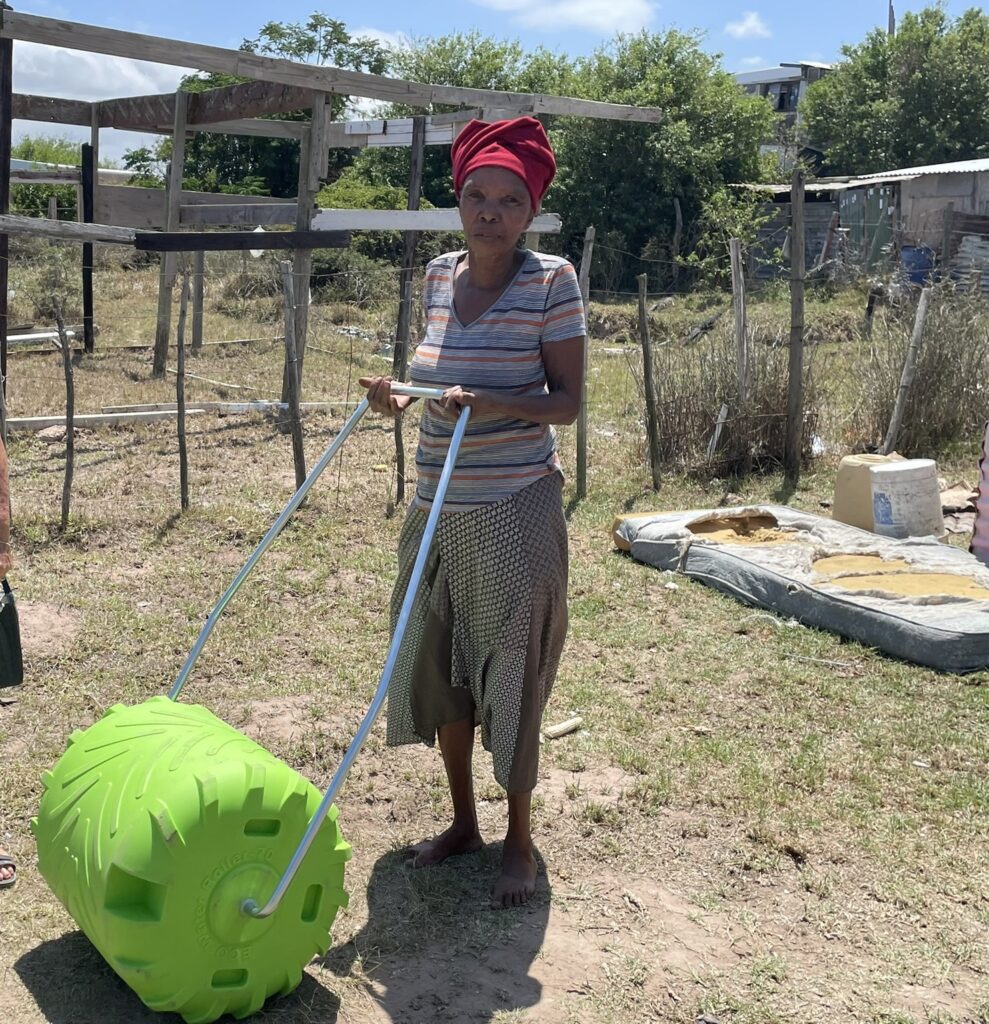 Woman in a headscarf pulls a bright green water roller across a grassy area, surrounded by simple structures and outdoor materials.