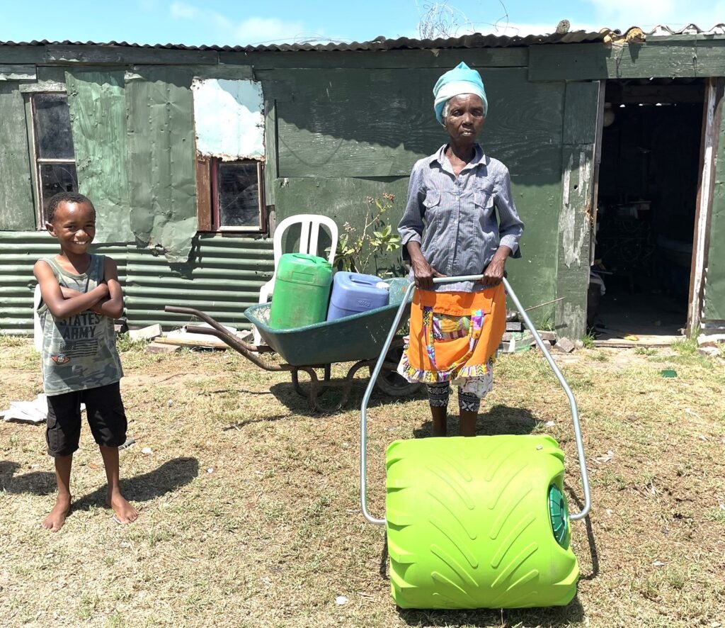 An elderly woman in a colorful apron stands holding a green wheelbarrow, while a young boy smiles beside her. They are outside a corrugated metal structure.