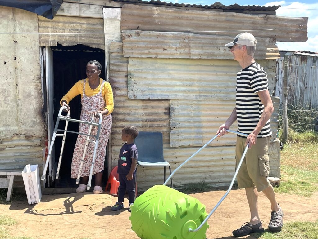 A woman exits a tin home with a walker, while a man pushes a green cart alongside a child. The scene highlights community support and care.