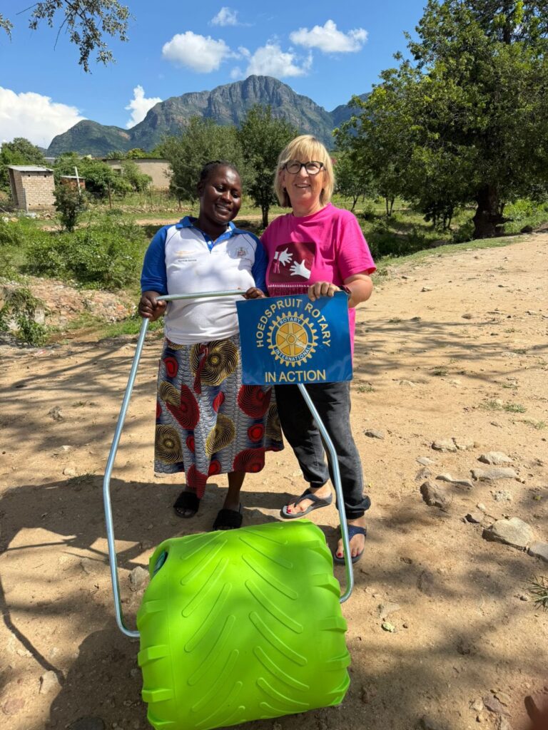 Two women, one holding a Rotary banner, stand beside a green water roller in a rural setting with mountains in the background. This highlights a community support initiative.
