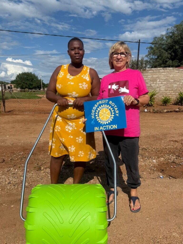 Two women stand together in a rural setting, one holding a sign that reads "Hoedspruit Rotary in Action" beside a green yard roller, symbolizing community support.