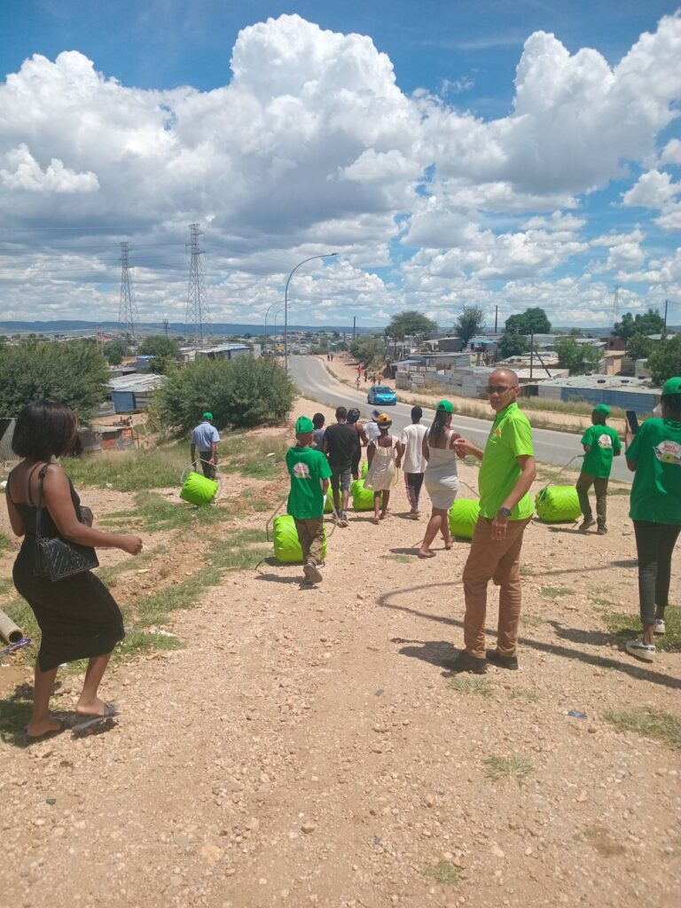 A group of people in green shirts and hats walks along a dirt path carrying bright green bags, under a cloudy sky. The scene suggests a community initiative.