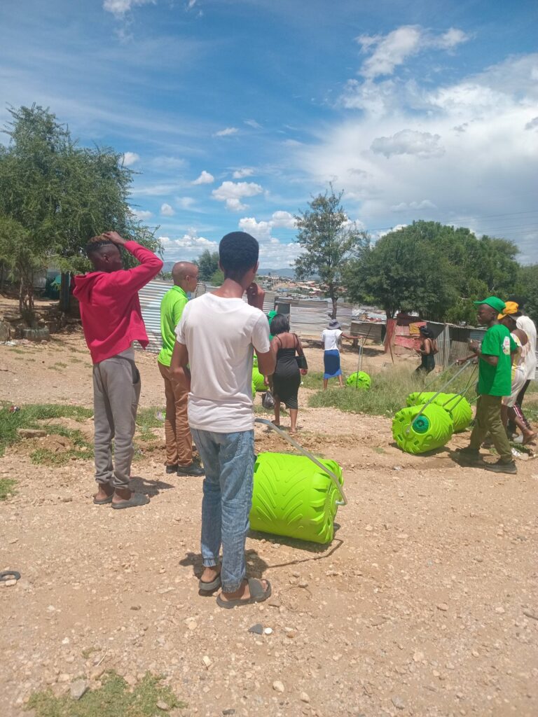 Youth in a community setting pull bright green rolling bins, likely part of a clean-up initiative, under a sunny sky.