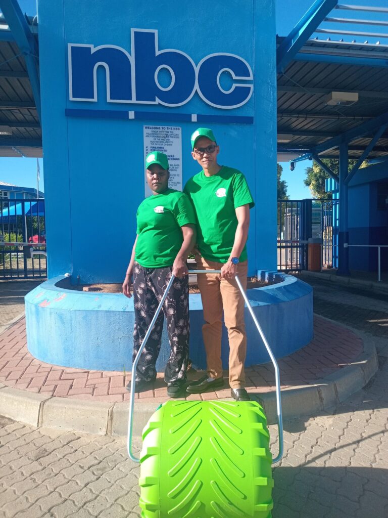 Two individuals in green shirts and caps stand beside a large, bright green roller near the NBC sign, promoting environmental awareness or clean-up efforts.