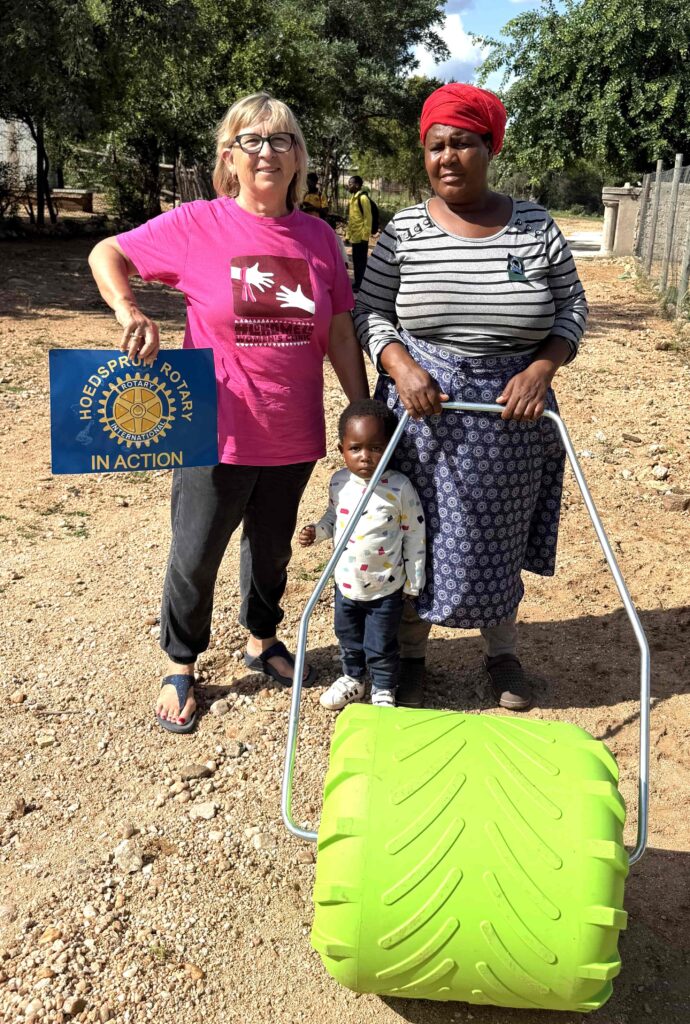 Two women stand beside a bright green gardening tool, with a child in a white jacket between them. One woman holds a sign for Hoedspruit Rotary, showcasing community support.